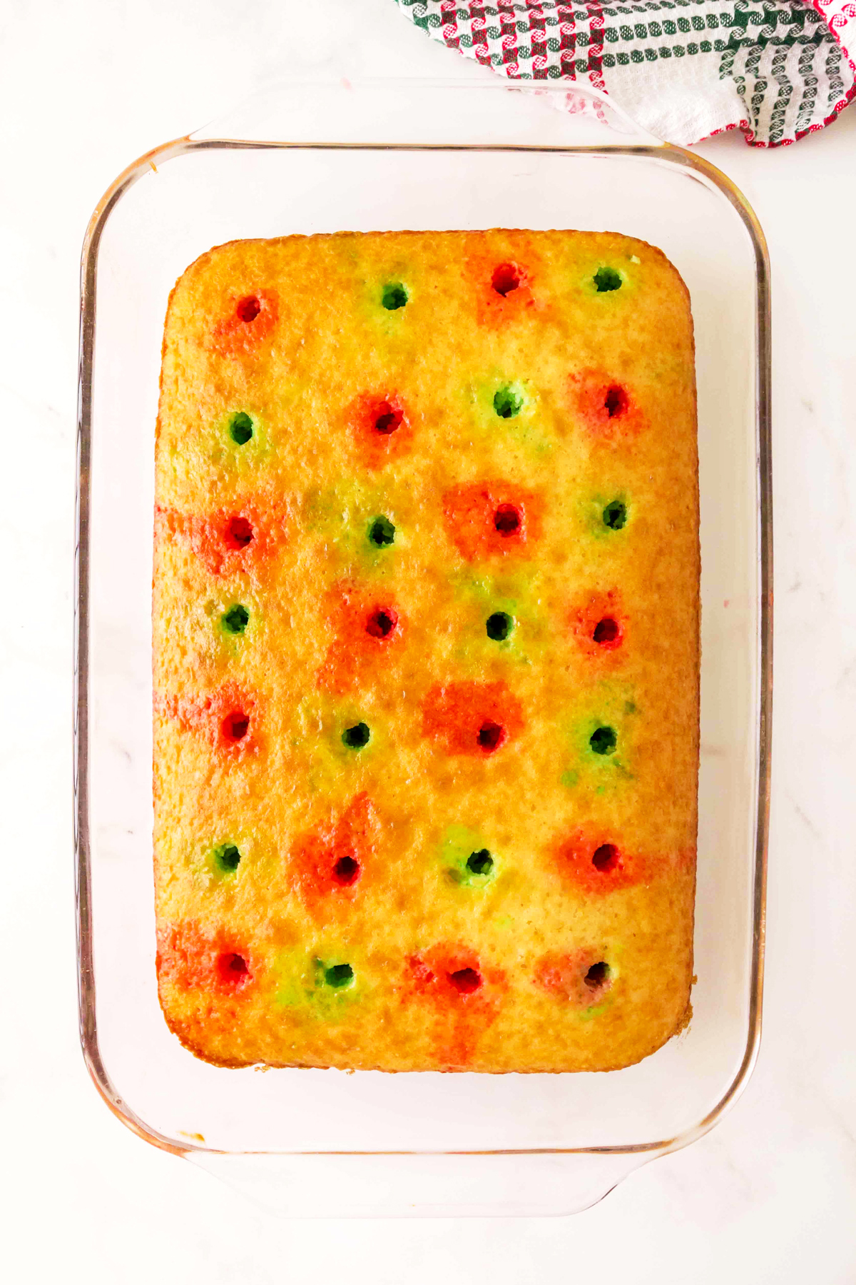 A rectangular Christmas Poke Cake in a glass baking dish with evenly spaced red and green holes on the surface. A textured cloth is partially visible in the top right corner.