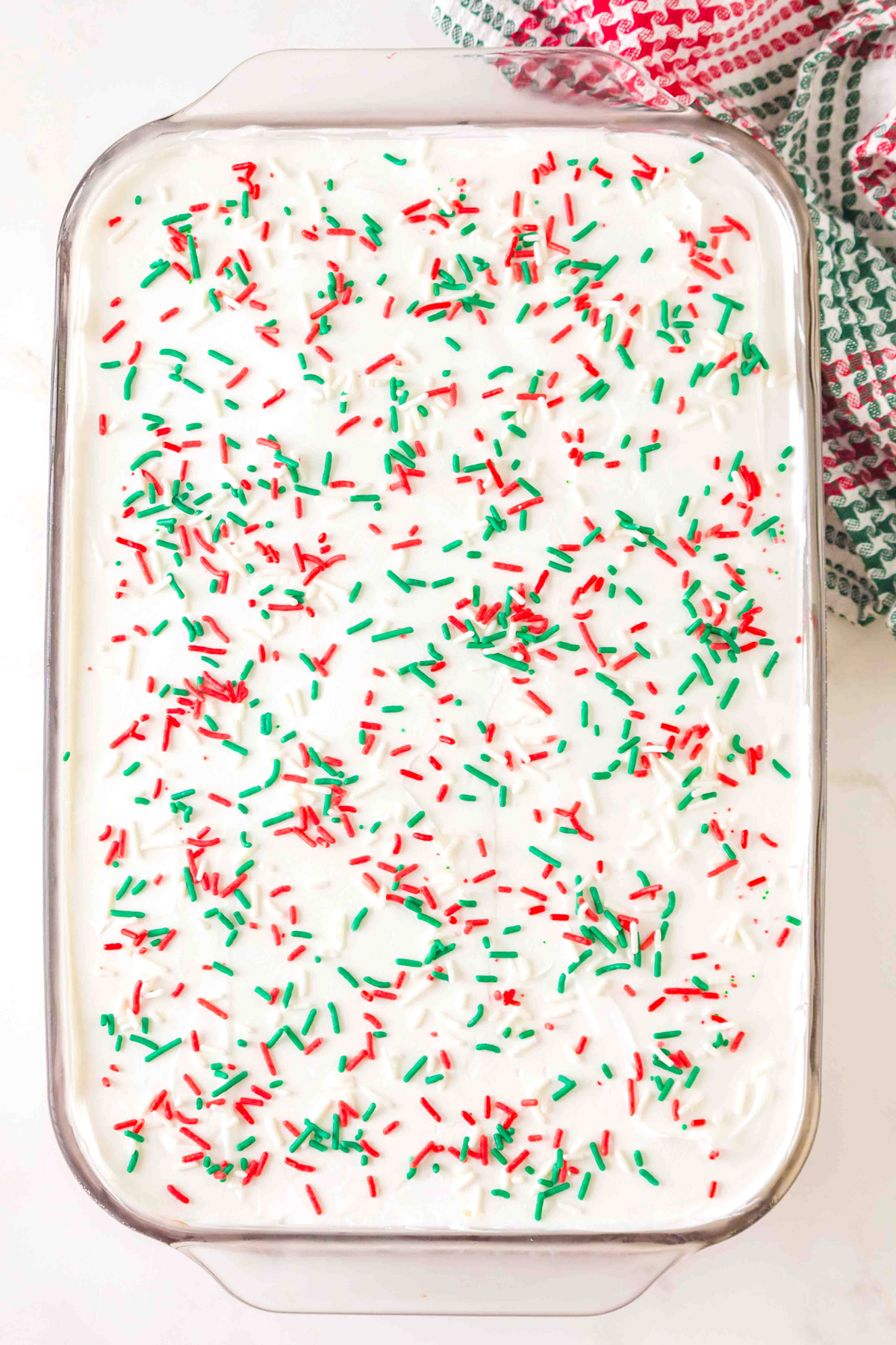 A rectangular glass baking dish filled with a dessert topped with white frosting and red and green sprinkles.