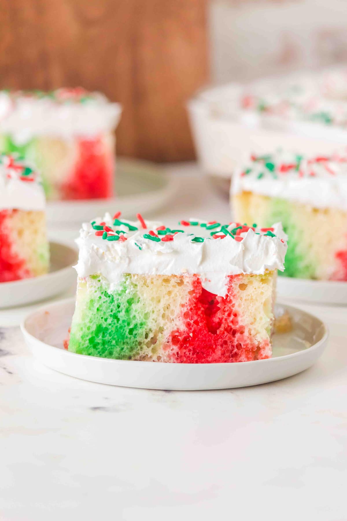 A slice of Christmas Poke Cake with red and green swirls, topped with white frosting and red and green sprinkles, on a white plate.