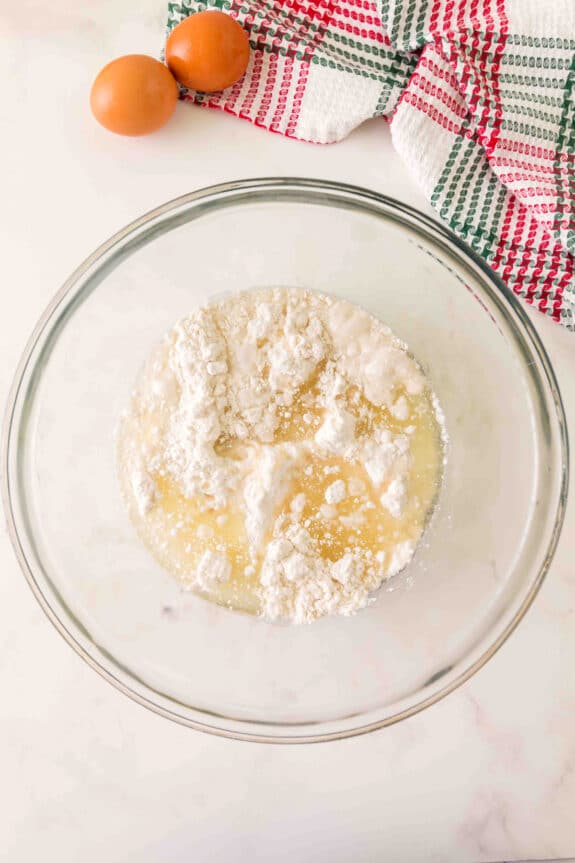 A glass bowl containing flour, liquid, and other baking ingredients sits on a countertop near two eggs and a checkered kitchen towel for Christmas Poke Cake
