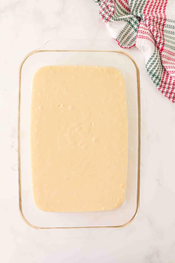 A glass baking dish filled cake mix batter for Christmas Poke Cake sits on a white surface beside a red, green, and white checkered cloth.