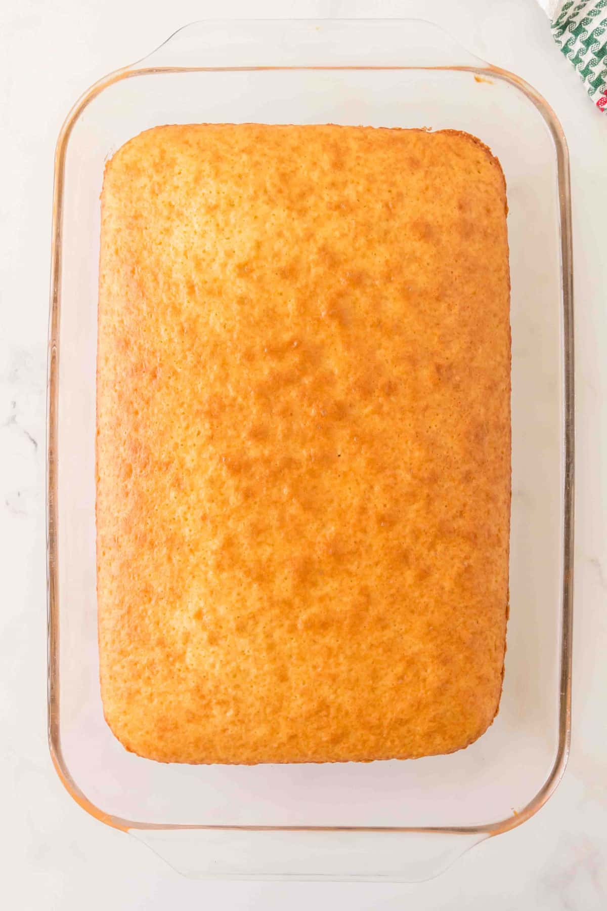 A rectangular, golden-brown baked cake sits in a clear glass baking dish on a white surface for Christmas Poke Cake recipe.