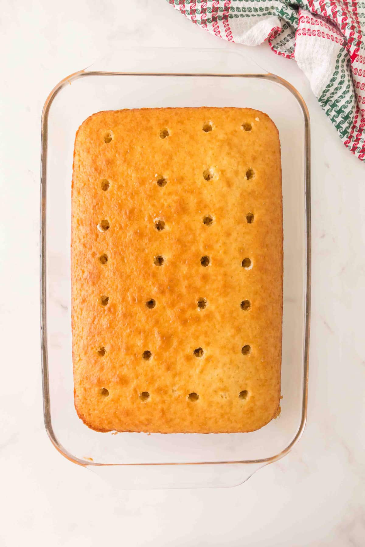 A rectangular sheet cake with evenly spaced holes for Christmas Poke Cake on top sits in a glass baking dish; a patterned kitchen towel is nearby.