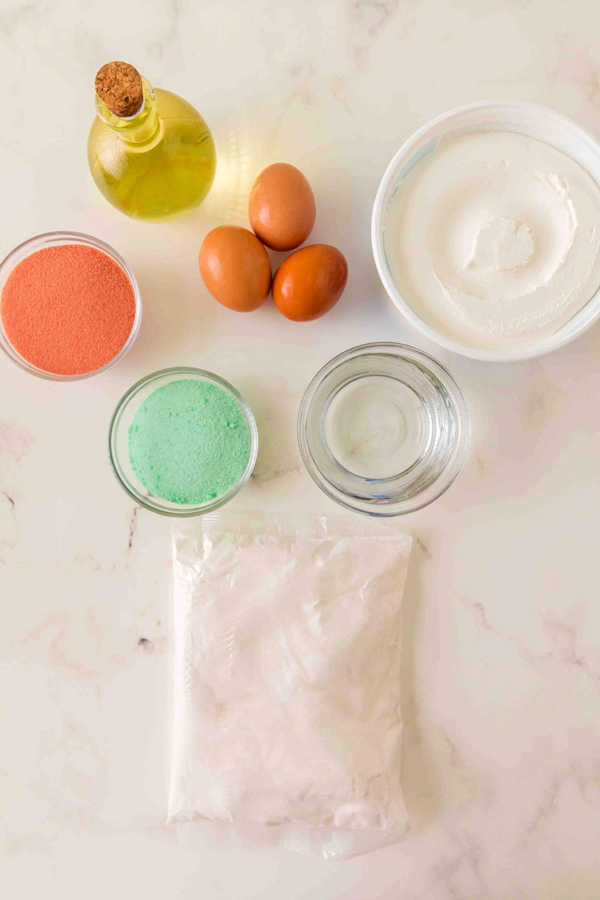 Ingredients for Christmas Poke Cake on a white marble counter are the following: white cake mix, water, oil, eggs, lime and strawberry Jell-O, boiling water, Cool Whip, red and green sprinkles.