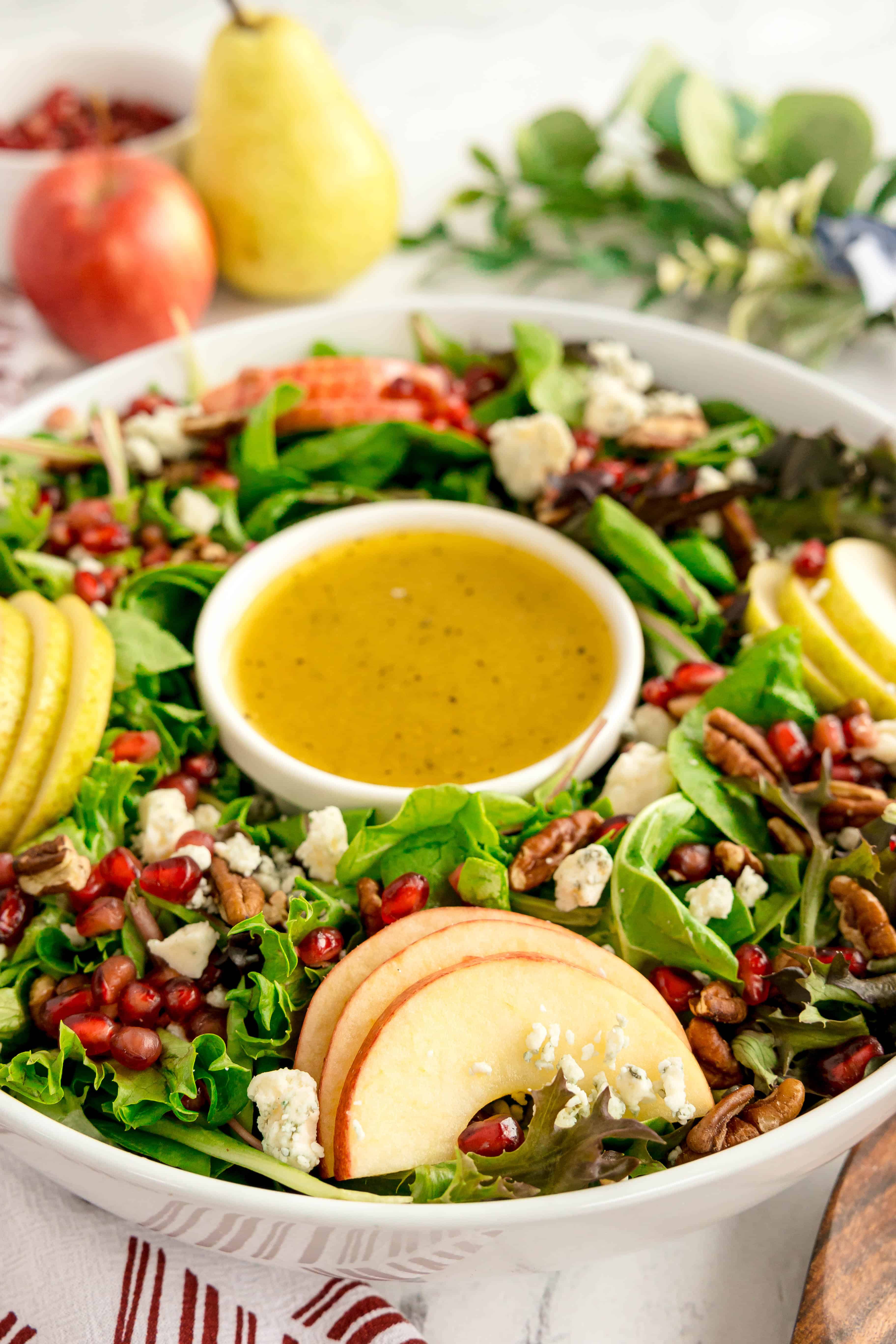 A festive Christmas wreath salad featuring mixed greens, apple and pear slices, pecans, pomegranate seeds, and blue cheese, beautifully arranged around a bowl of yellow vinaigrette dressing.