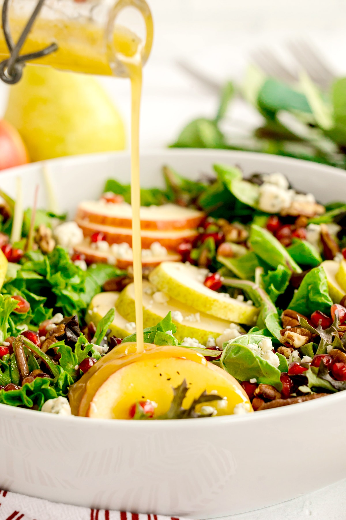 A close-up of a Christmas Wreath Salad with pear slices, pomegranate seeds, leafy greens, nuts, and crumbled cheese, as dressing is poured over it from a small glass container.