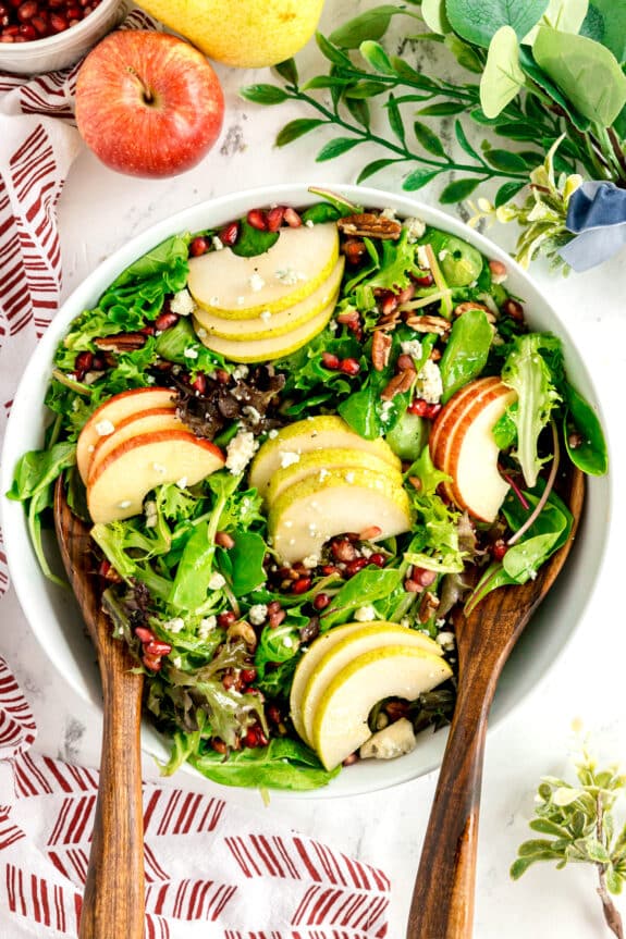 A bowl of Christmas Wreath Salad topped with sliced apples, pears, pomegranate seeds, pecans, and cheese, with wooden salad servers on each side.
