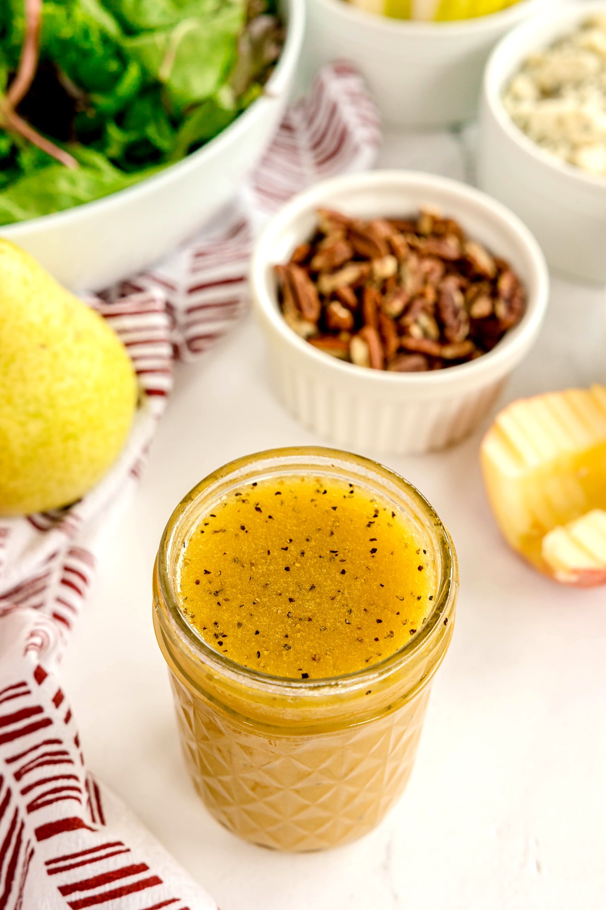 A jar of vinaigrette dressing sits on a counter near pecans, a pear, an apple slice, leafy greens, and a bowl of blue cheese—perfect ingredients for creating a festive Christmas wreath salad.