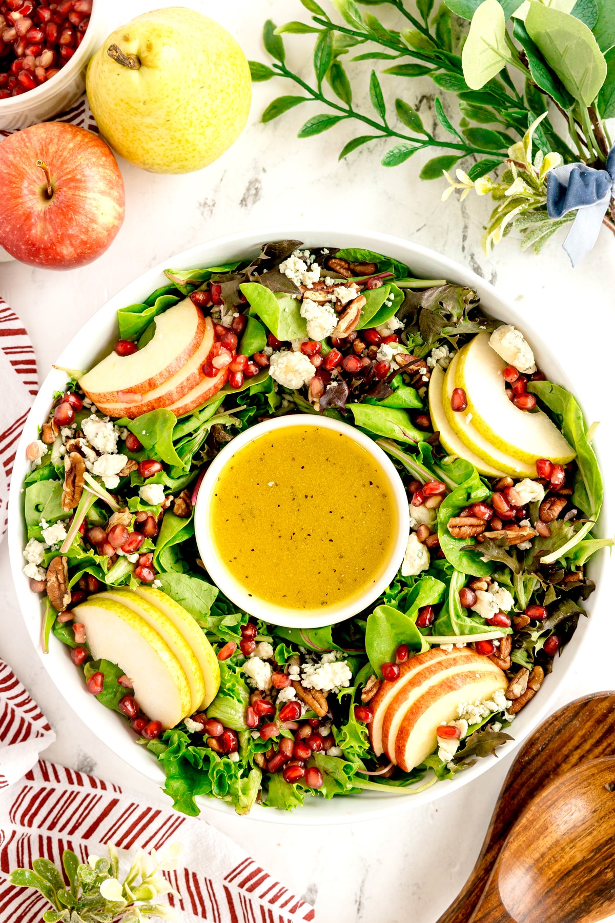 A bowl of Christmas Wreath Salad topped with apple slices, crumbled cheese, pecans, and pomegranate seeds, with a small bowl of dressing in the center. Apples and greens are nearby.