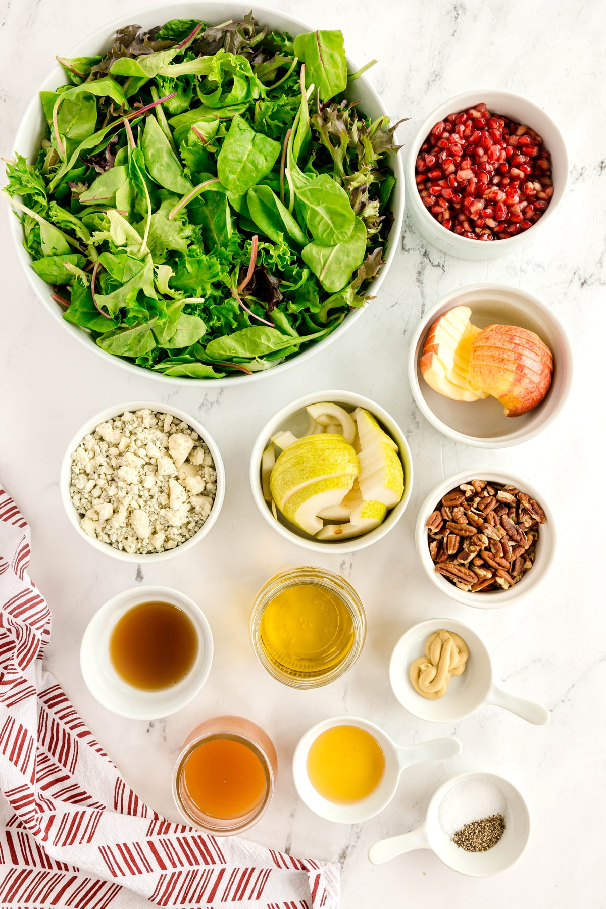 Ingredients for Christmas Wreath Salad are the following: a bowl of mixed greens, pomegranate seeds, apple slices, blue cheese, pecans, and ingredients for dressing such as olive oil, apple cider vinegar, dijon mustard, honey and maple syrup, salt and black pepper, are arranged on a white surface with a red and white napkin.