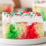 A slice of Christmas poke cake with red and green swirls, topped with white frosting and festive sprinkles, sits on a white plate.