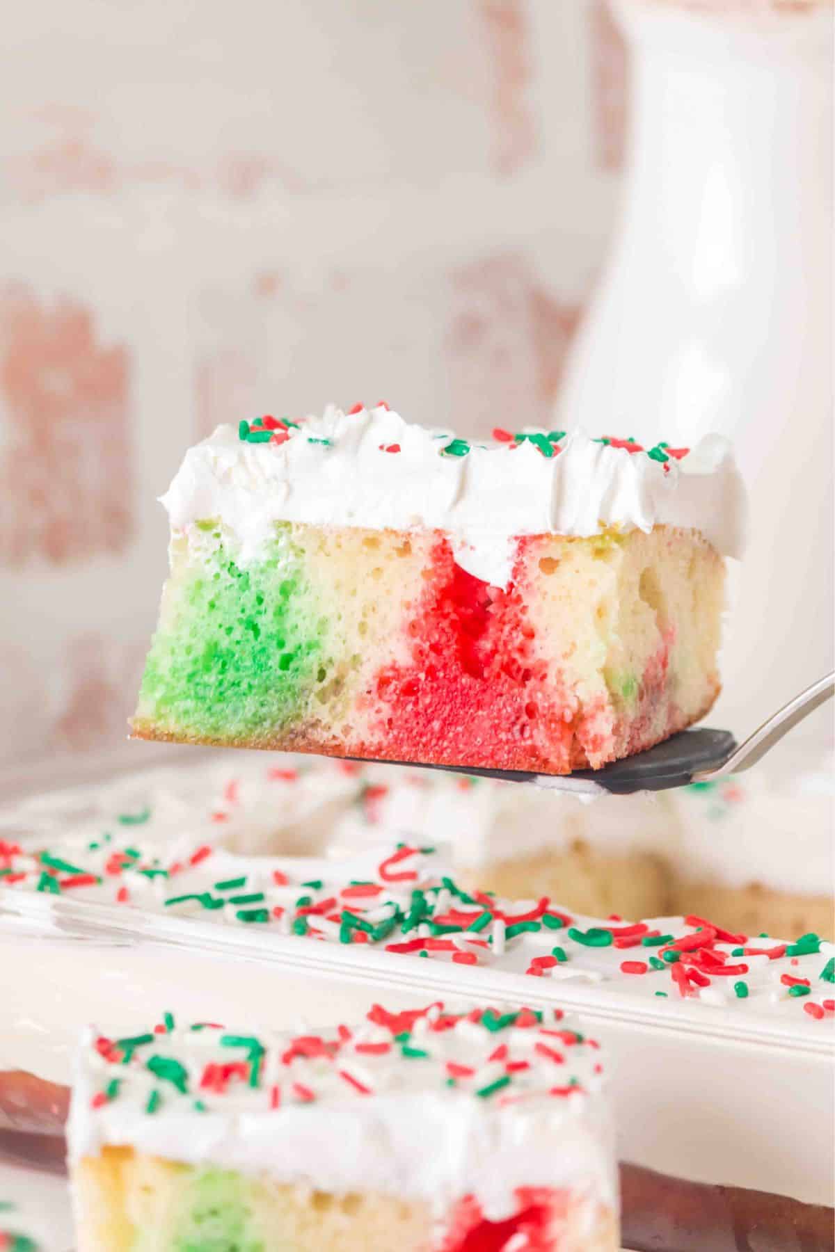A square slice of Christmas poke cake with red and green colors, topped with white frosting and festive sprinkles, is being lifted from a cake pan.