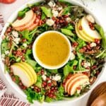 A festive christmas wreath salad featuring mixed greens, sliced apples, pecans, pomegranate seeds, blue cheese crumbles, and a bowl of dressing in the center.