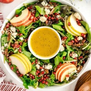 A festive christmas wreath salad featuring mixed greens, sliced apples, pecans, pomegranate seeds, blue cheese crumbles, and a bowl of dressing in the center.