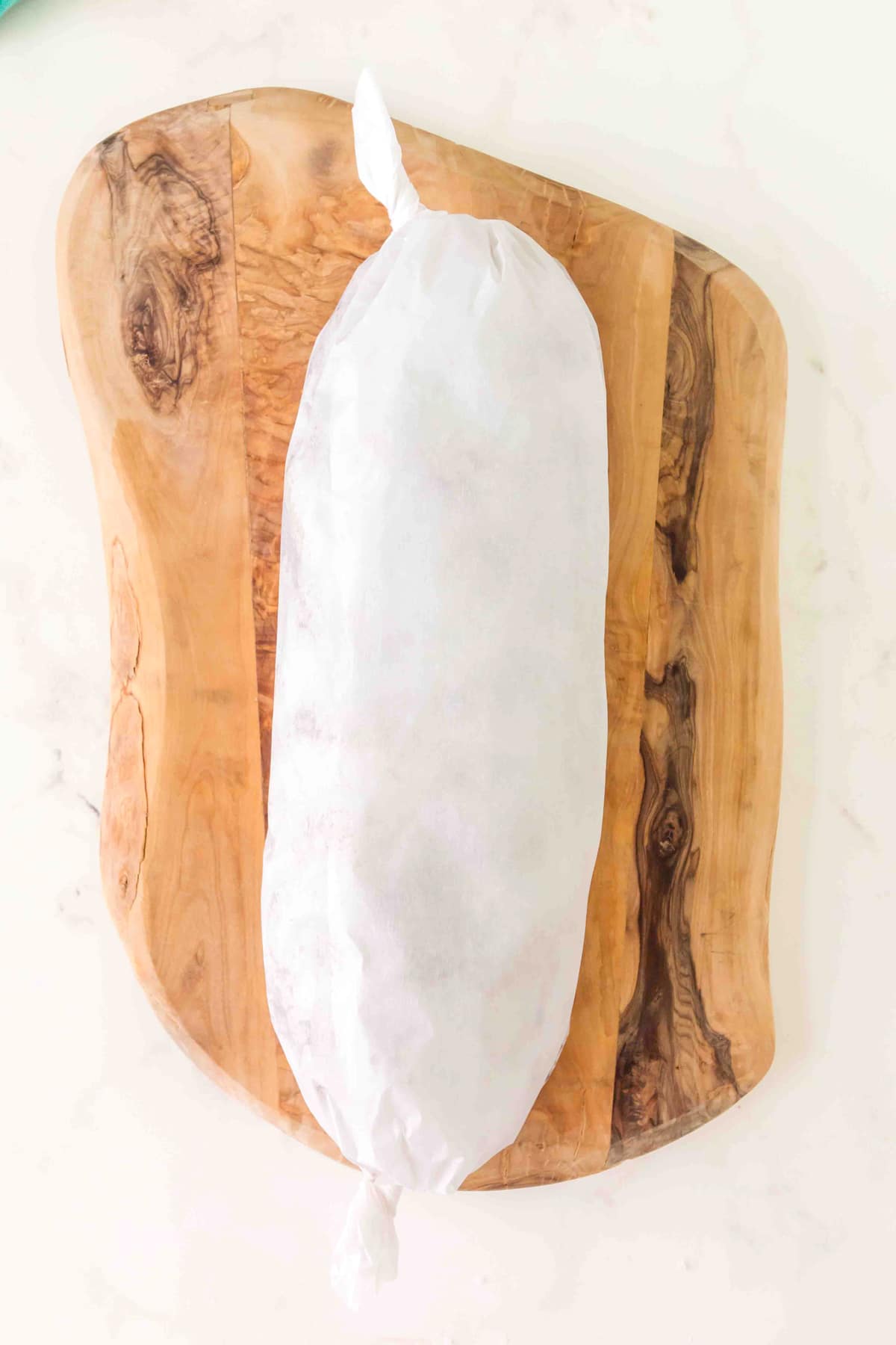 A wrapped Church Window Cookies, secured at both ends, sits on a wooden cutting board placed on a light surface, ready to be put in the fridge.