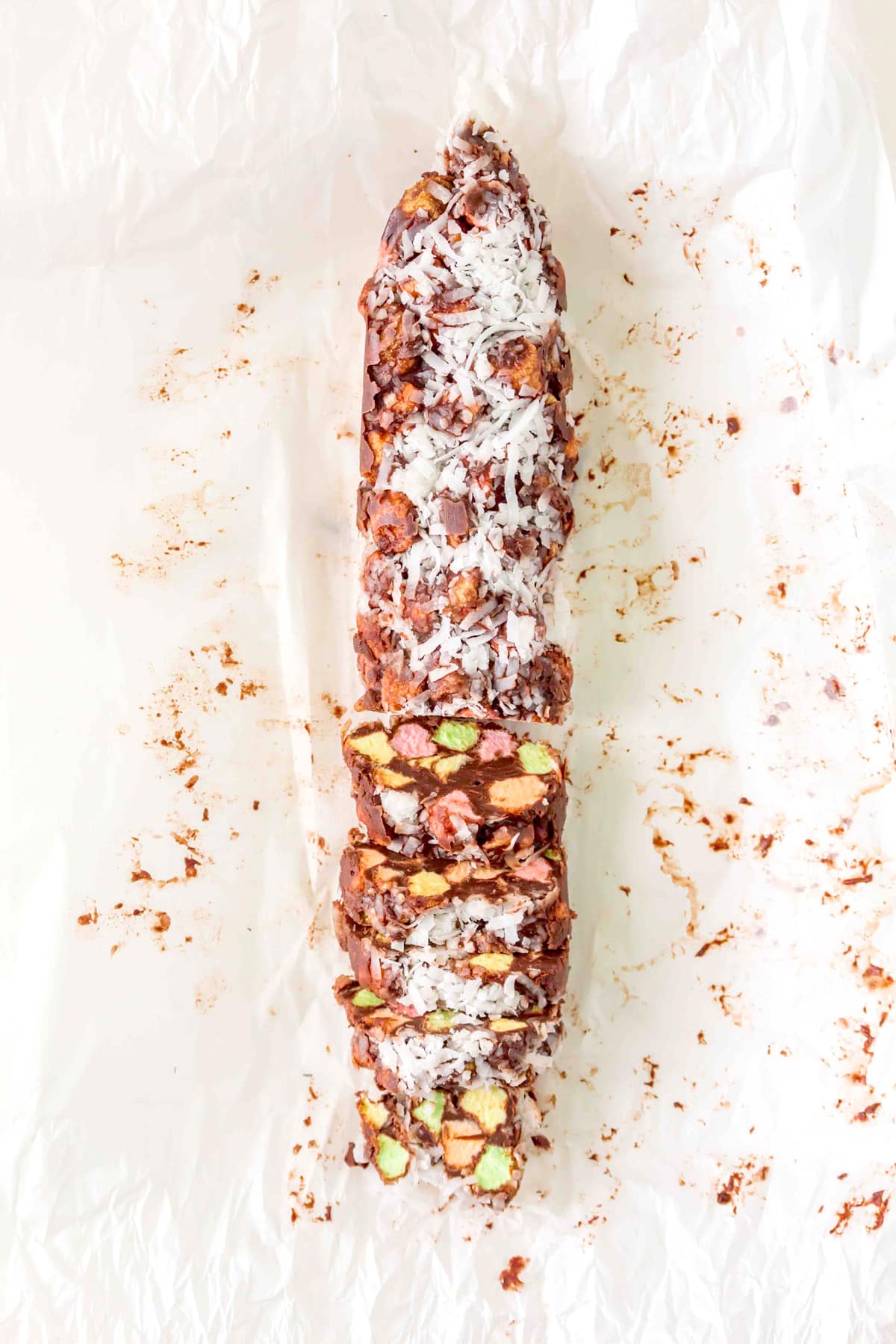 A Church Window Cookies log topped with shredded coconut is sliced to reveal colorful pieces inside, resting on crumpled parchment paper.