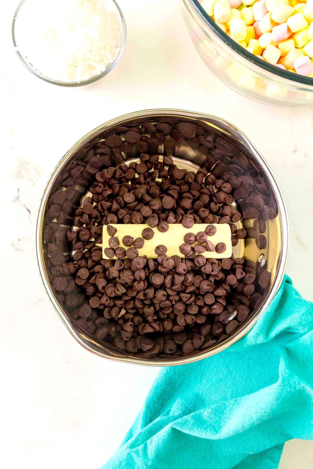 First step in preparing Church Window Cookies is to use a metal bowl filled with chocolate chips and a stick of butter, with bowls of shredded coconut and colorful marshmallows nearby on a white surface.