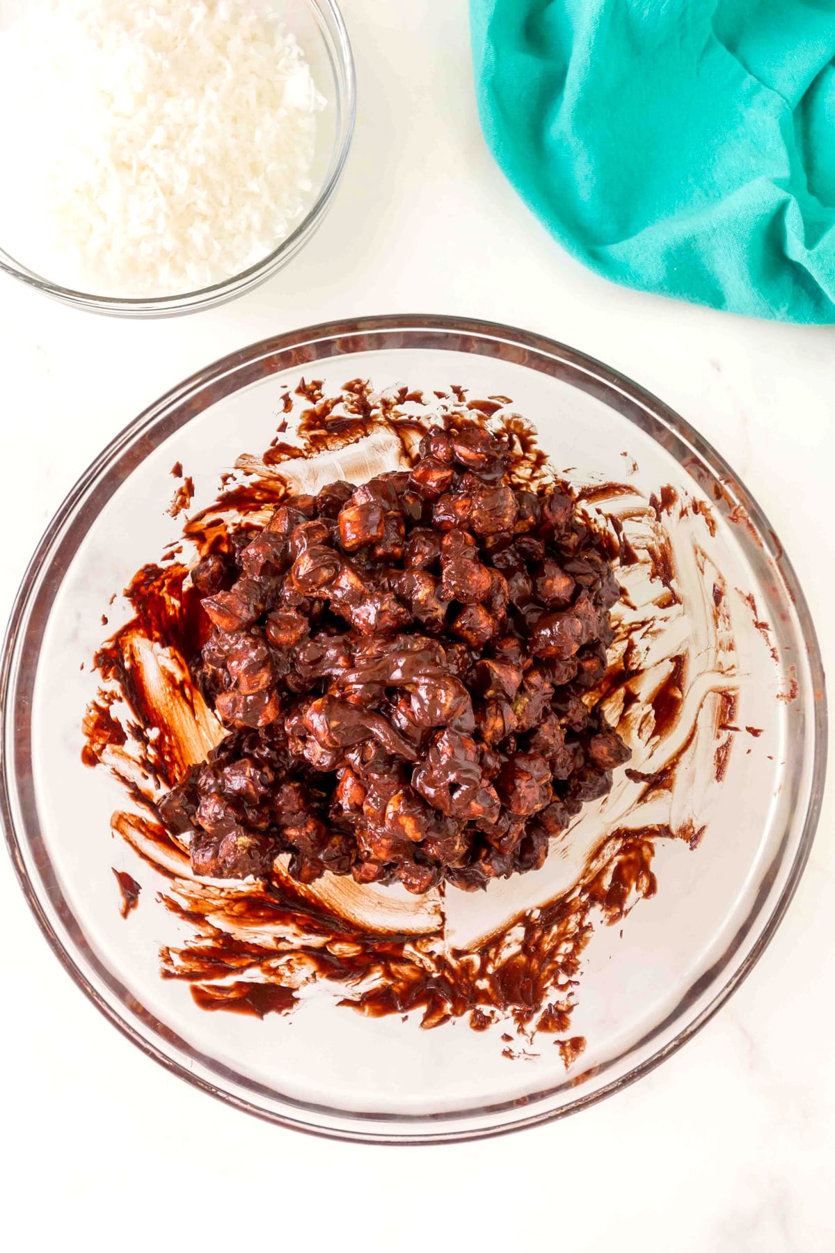 A glass bowl filled with a mixture of melted chocolate and marshmallows sits on a white surface next to a bowl of shredded coconut and a teal cloth.