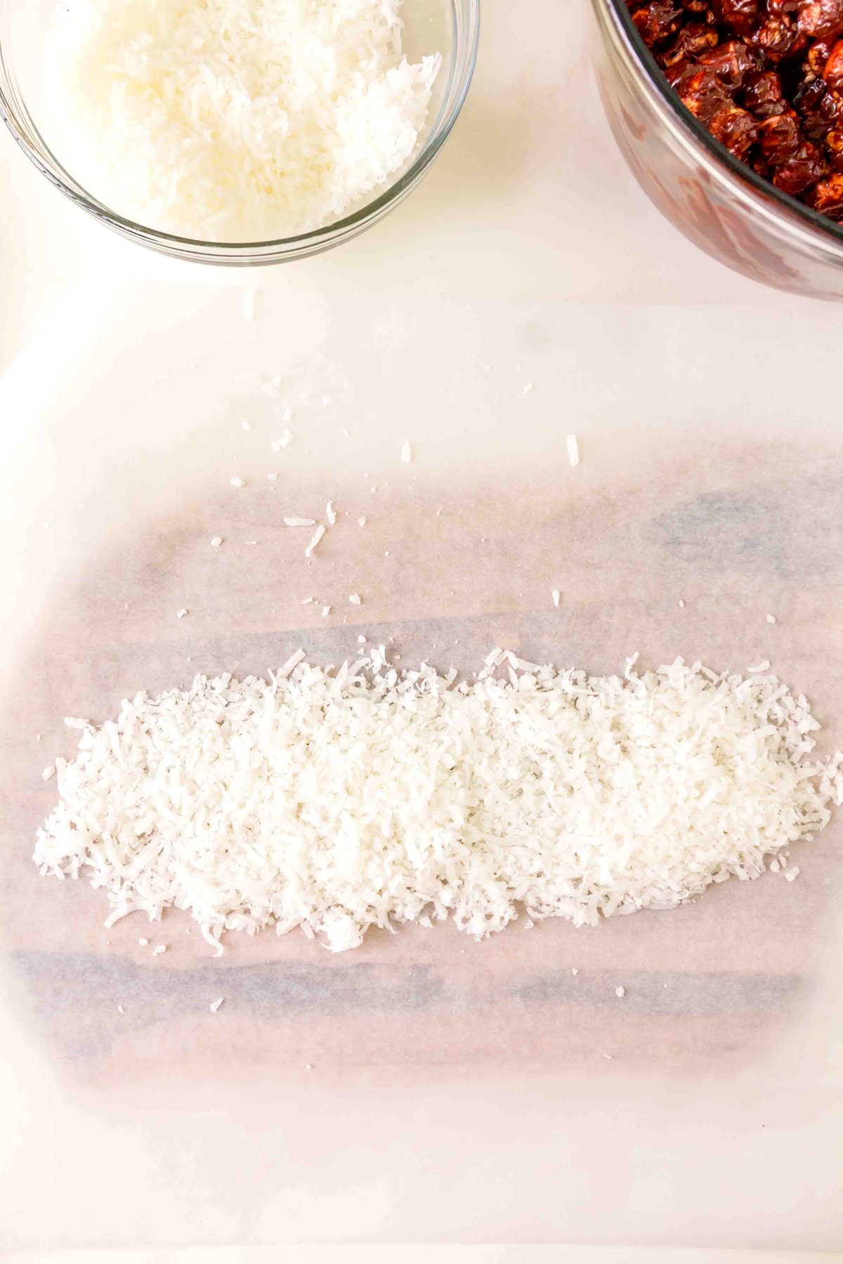 A line of shredded coconut is spread on parchment paper, with a bowl of shredded coconut and a bowl of chopped dates nearby.