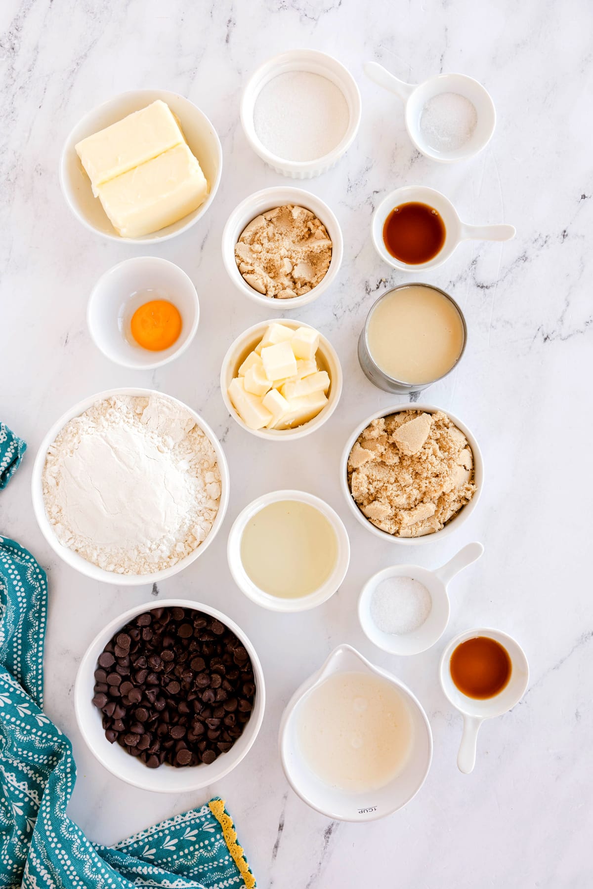 Assorted baking ingredients for Millionaire Shortbread Bars in small bowls on a marble surface, including butter, sugar, all-purpose flour, egg, chocolate chips, vanilla extract, light corn syrup, salt, brown sugar, whipping cream, and liquids, with a blue patterned cloth in the corner.