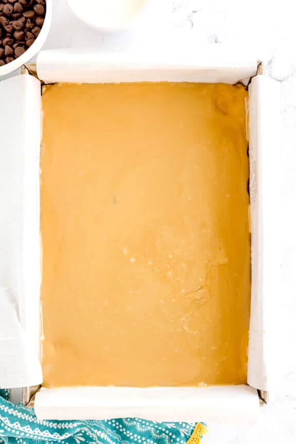 A square baking pan lined with parchment paper is filled with a smooth layer of caramel mixture for Millionaire Shortbread Bars; bowls with ingredients are partially visible nearby.