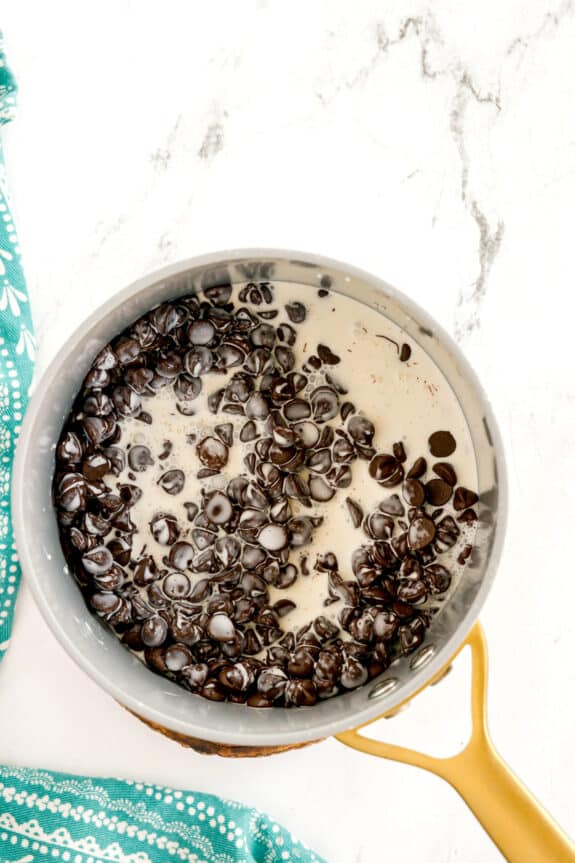Next step is preparing the Chocolate Ganache Layer for Millionaire Shortbread Bars A saucepan with chocolate chips and cream, partially melted and mixed, sits on a white marble surface next to a blue patterned cloth.