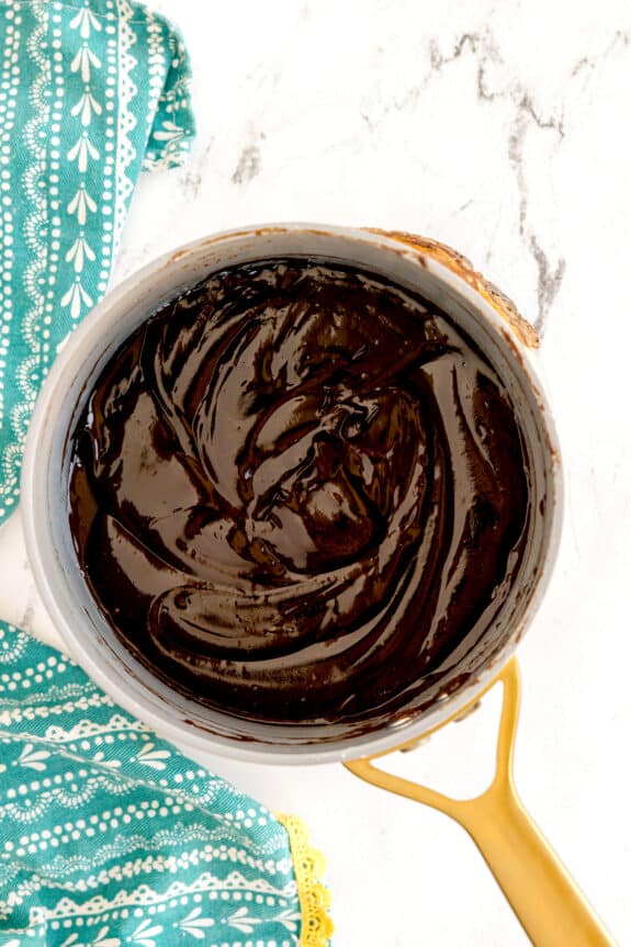 A saucepan filled with thick, glossy chocolate mixture for Millionaire Shortbread Bars sits on a white marble surface next to a green patterned cloth.