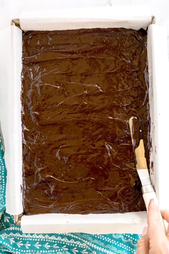 A hand uses an offset spatula to spread chocolate batter for Millionaire Shortbread Bars, evenly in a parchment-lined rectangular baking pan.
