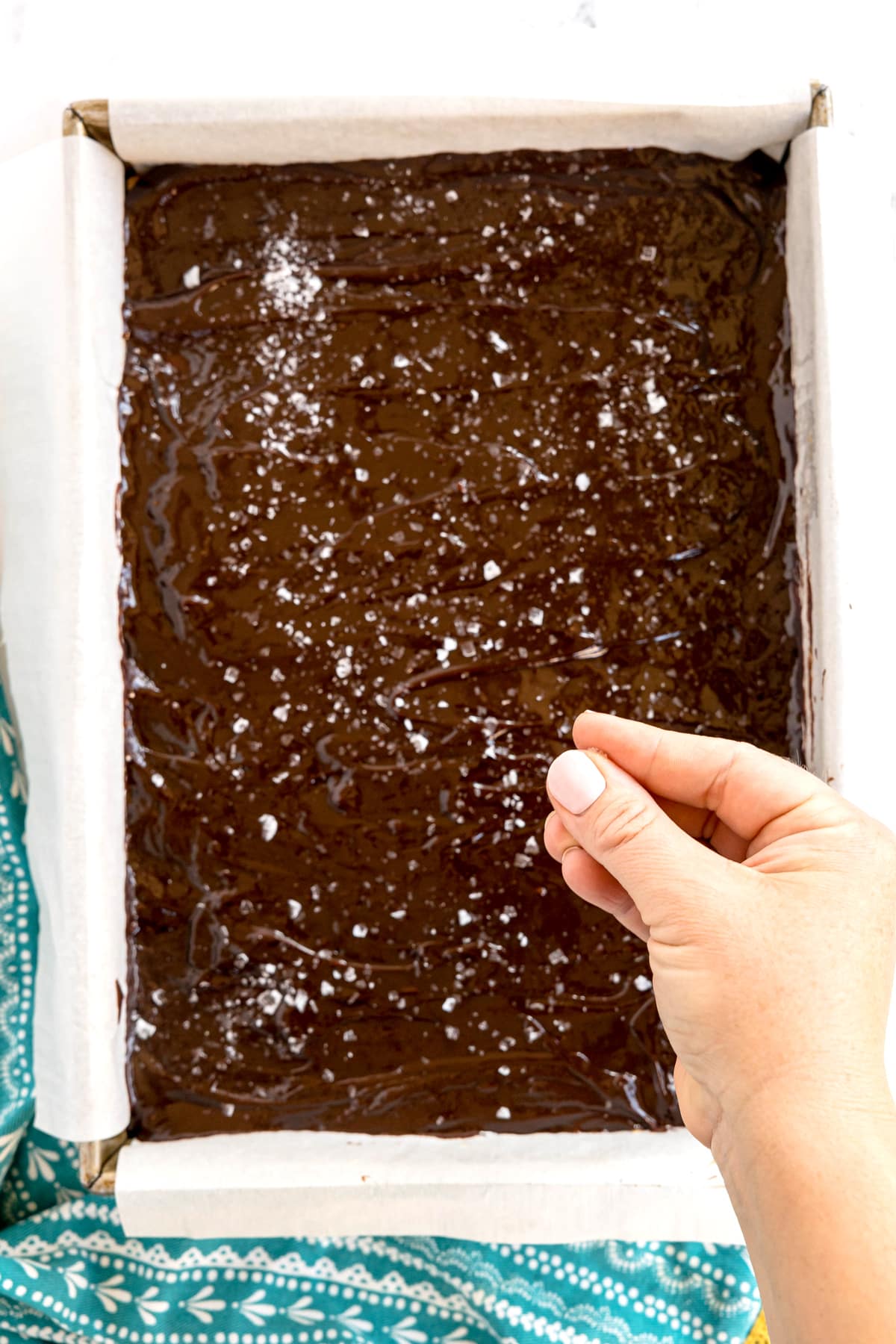 A hand sprinkles sea salt over a rectangular pan of Millionaire Shortbread Bars lined with parchment paper, set on a blue patterned cloth.