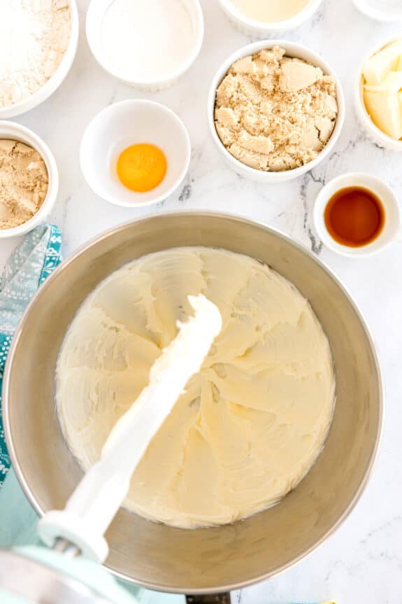 Overhead view of a mixing bowl with creamed butter, surrounded by ingredients in bowls including brown sugar, an egg yolk, flour, sugar, vanilla extract, and butter on a marble surface for Millionaire Shortbread Bars recipe