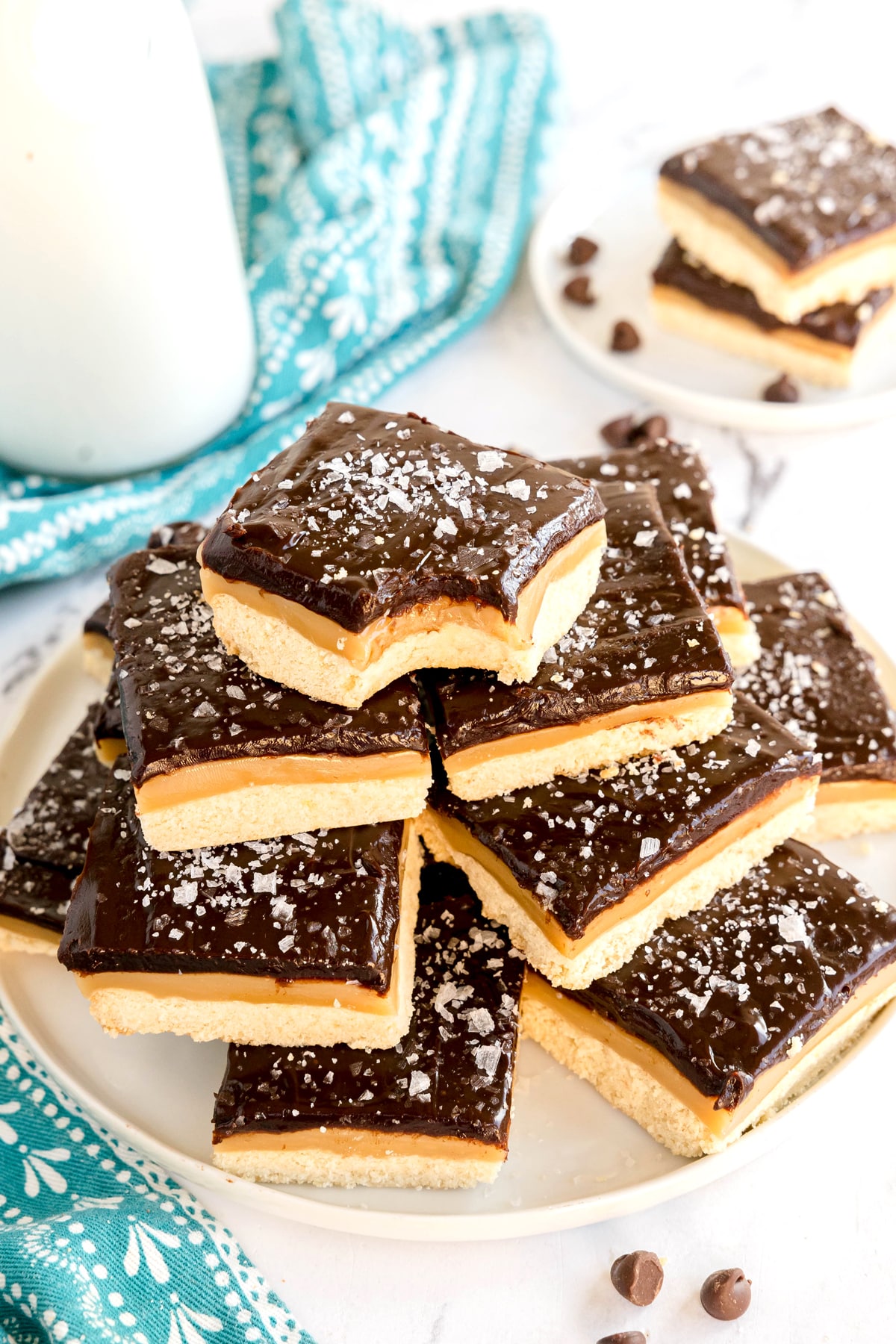 A plate of stacked Millionaire Shortbread Bars topped with flaky sea salt, with a blue patterned cloth and more bars in the background.