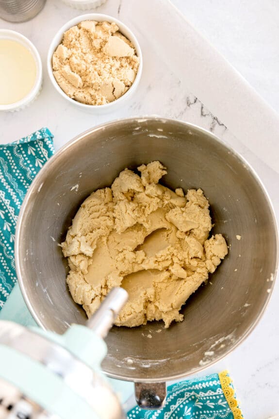 A mixing bowl filled with Millionaire Shortbread Bars dough sits on a counter next to a small bowl of brown sugar and a cup of liquid.