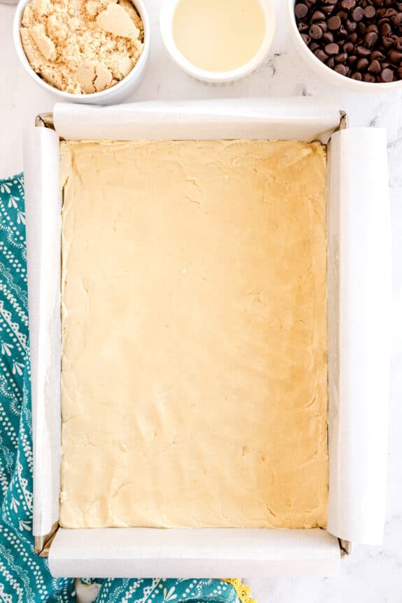 A square baking pan lined with parchment paper contains an unbaked Millionaire Shortbread Bars dough. Nearby are bowls of brown sugar, a yellow liquid, and chocolate chips. A teal cloth is partially visible.