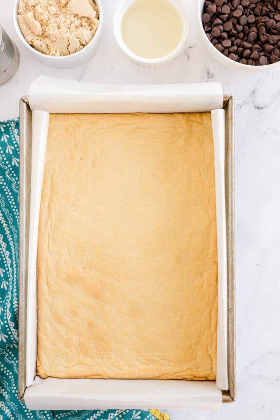 A baked Millionaire Shortbread Bars dough in a parchment-lined pan, with bowls of brown sugar, vegetable oil, and chocolate chips nearby on a marble surface.