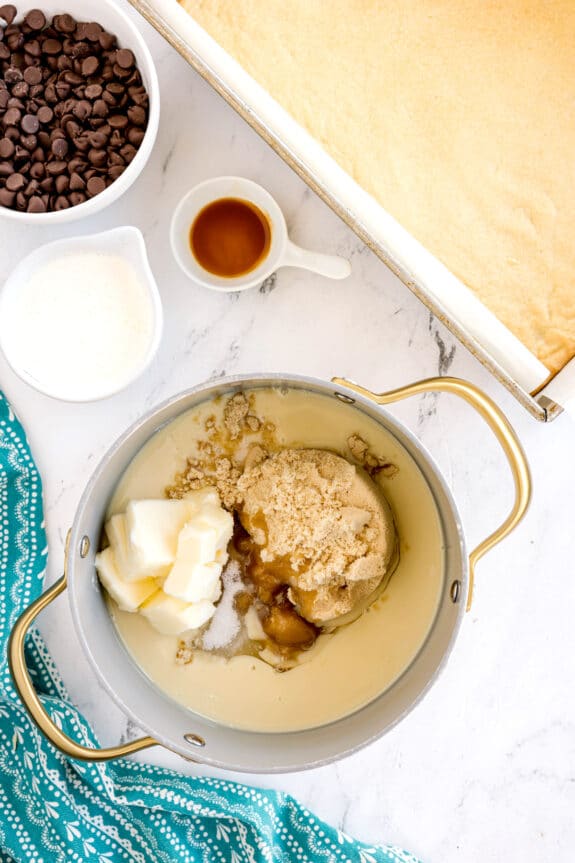 Next step in preparing Millionaire Shortbread Bars is to use a saucepan with sweetened condensed milk, butter, brown sugar, corn syrup, and salt. On a counter next to a bowl of chocolate chips, a bowl of cream, a cup of vanilla, and a baked crust in a baking tray can be seen.