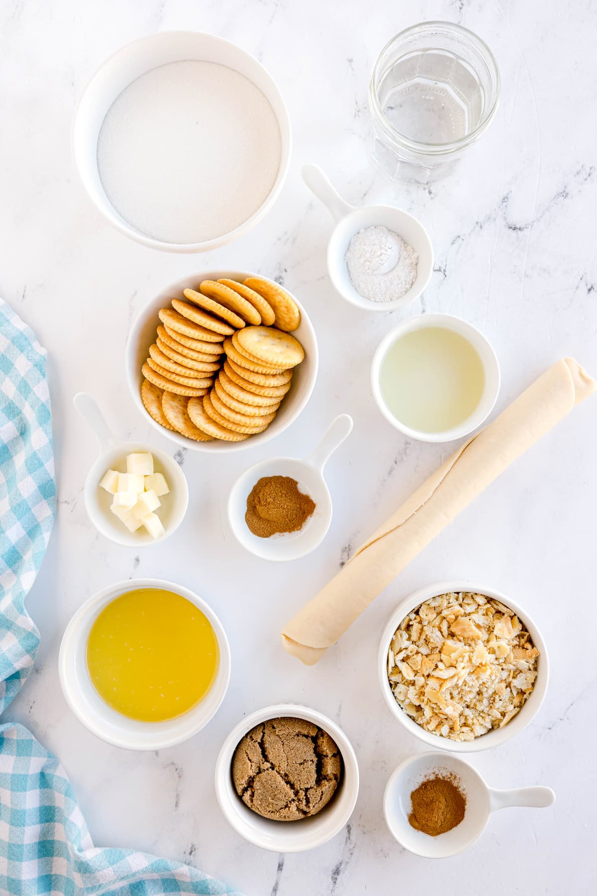 An assortment of baking ingredients for Ritz Cracker Pie on a white surface, including Ritz crackers, ganulated sugar, melted butter, brown sugar, unbaked 9-inch pie crust, ground cinnamon, and hot water.