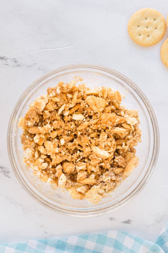 A glass bowl filled with a Ritz Cracker Pie mixture, likely crushed crackers and a brown ingredient, sits on a white surface next to a few whole crackers.