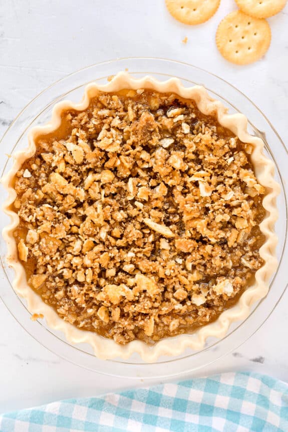 Uncooked Ritz Cracker Pie with a crimped crust and a crumbly brown topping sits on a white surface next to round crackers and a blue checkered cloth.