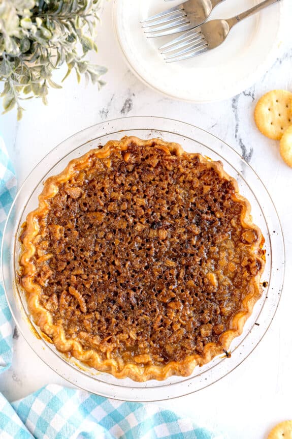 A baked Ritz Cracker Pie with a golden, crimped crust and a browned, textured filling sits in a glass pie dish on a white surface near a plate with forks, crackers, and a blue checked cloth.