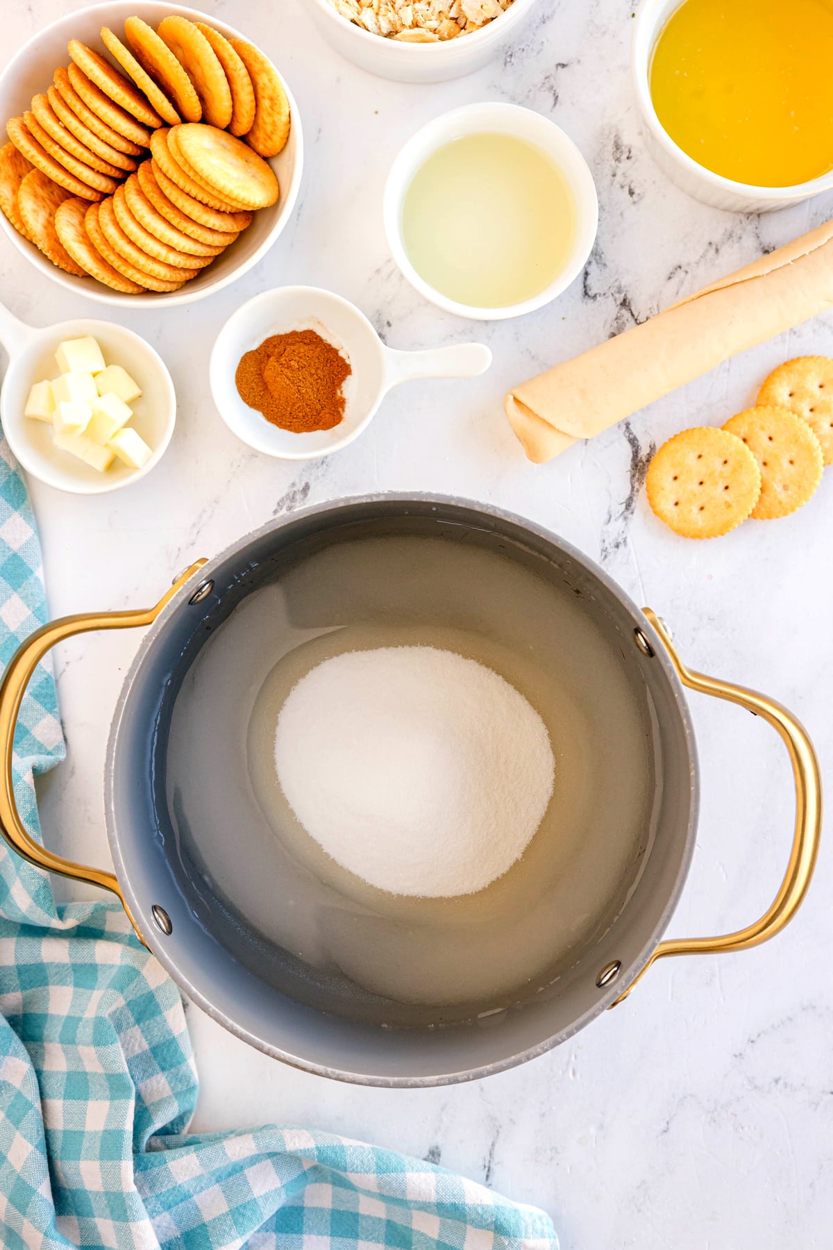 A medium saucepan containing hot water, granulated sugar, and cream of tartar. surrounded by bowls of crackers, butter, cinnamon, oats, oil, and a rolled pastry dough for Ritz Cracker Pie recipe