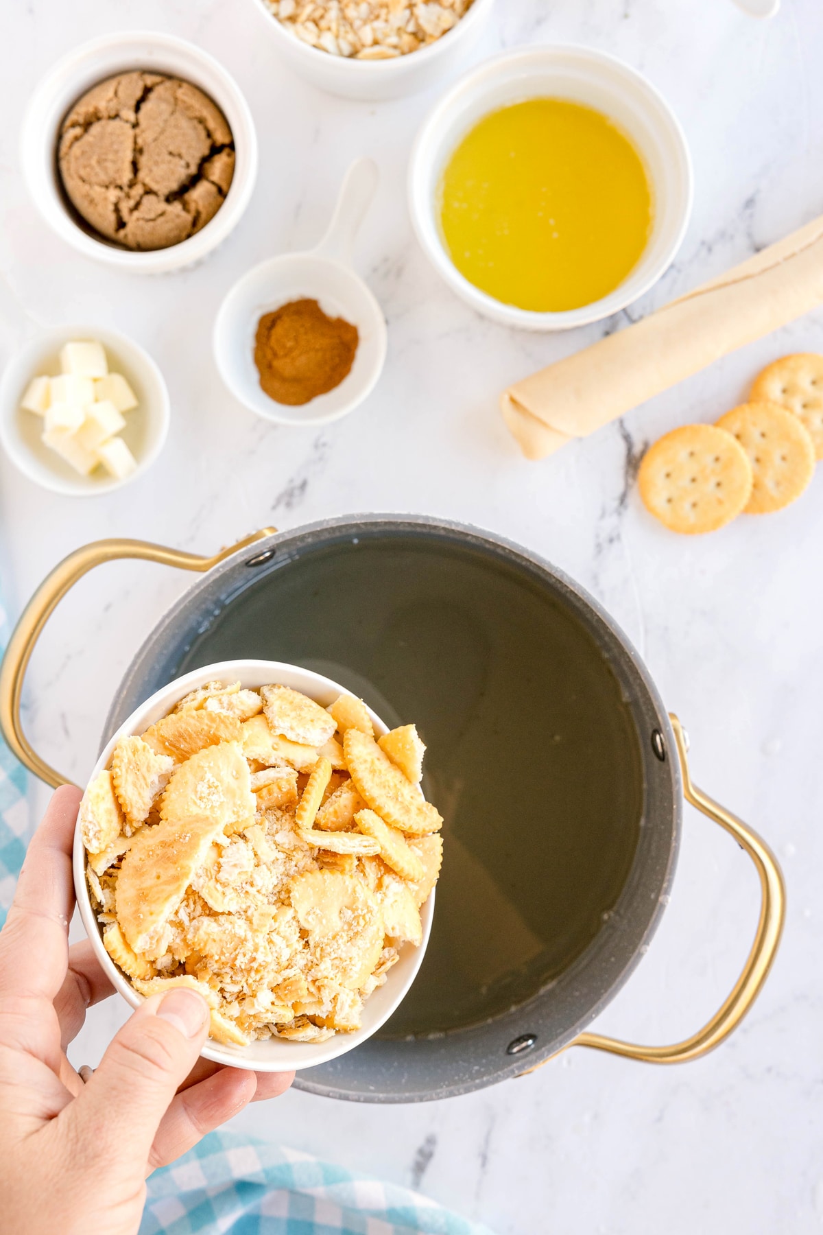 A hand holds a bowl of crushed crackers over a pot; surrounding the pot are bowls of brown sugar, cinnamon, melted butter, and cubed butter on a white surface. This is one step in preparing Ritz Cracker Pie