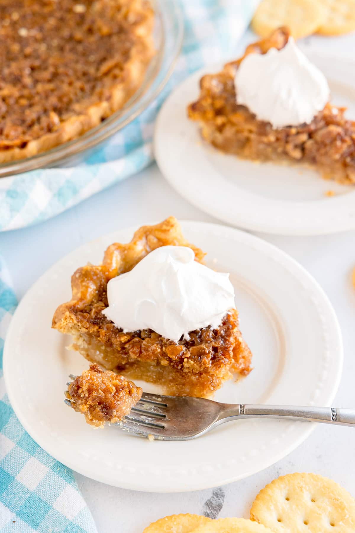 A slice of Ritz Cracker Pie with a dollop of whipped cream on a white plate, a fork holding a bite, and the rest of the pie and crackers in the background.