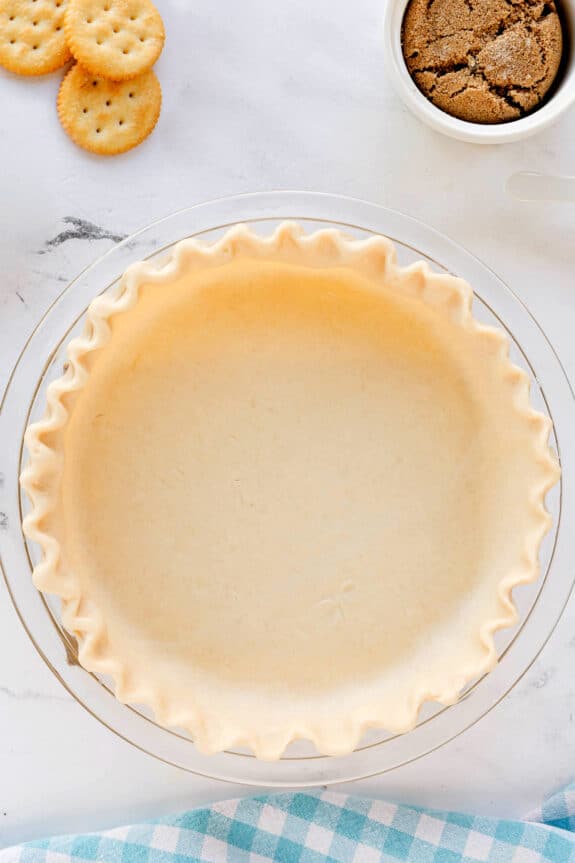 A raw pie crust in a glass dish for Ritz Cracker Pie, sits on a marble surface, with round crackers and a bowl of brown sugar nearby.