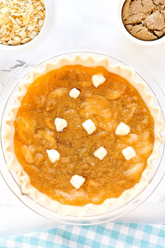 Unbaked Ritz Cracker Pie with a golden filling, sprinkled with brown sugar and topped with small cubes of butter. Bowls of crushed crackers and brown sugar are nearby.