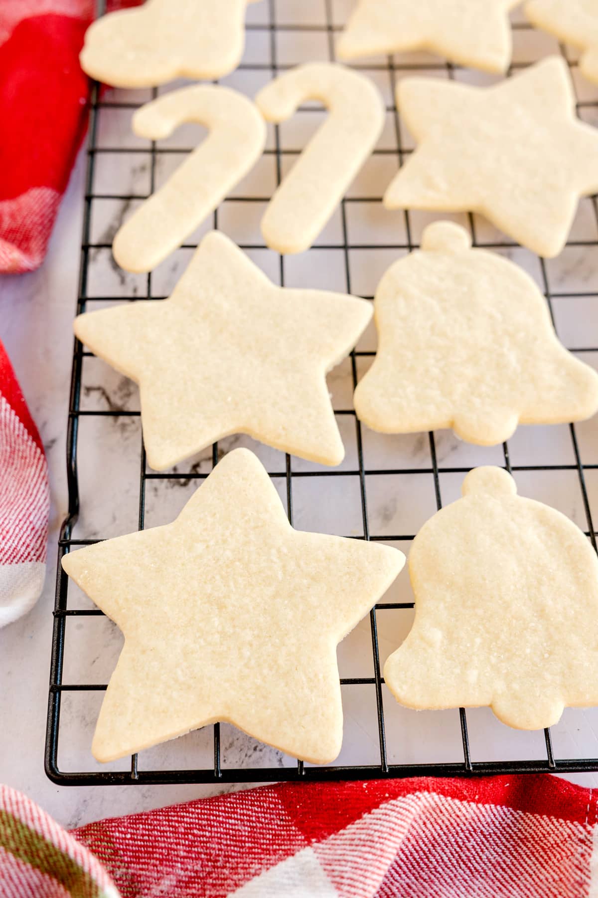 Unbaked Christmas Sugar Cookies in various shapes, including stars, bells, and candy canes, are arranged on a black cooling rack over a red and white cloth.