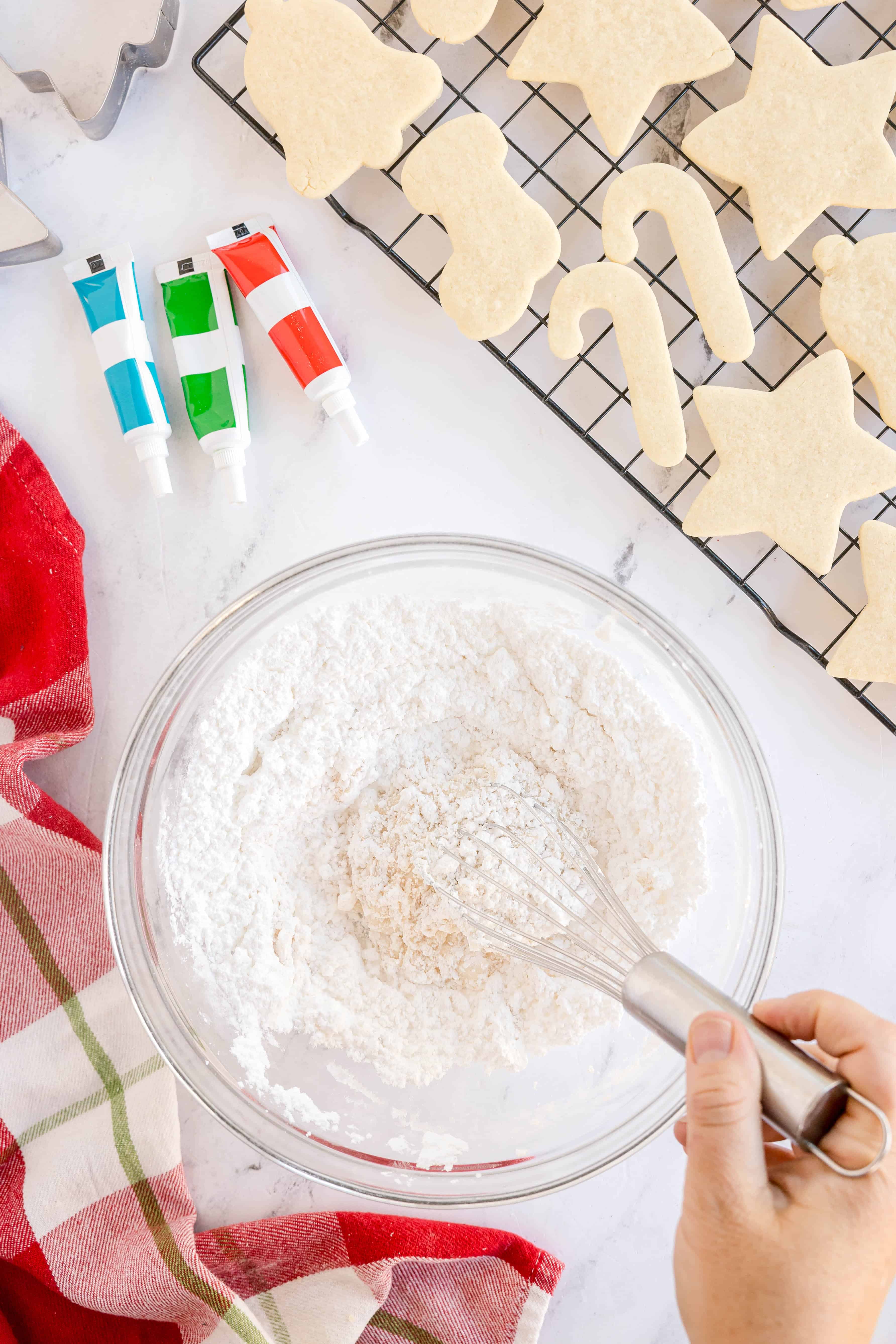 A hand whisks powdered sugar in a glass bowl next to a red and green towel, icing tubes, and unbaked holiday-shaped cookies on a cooling rack.