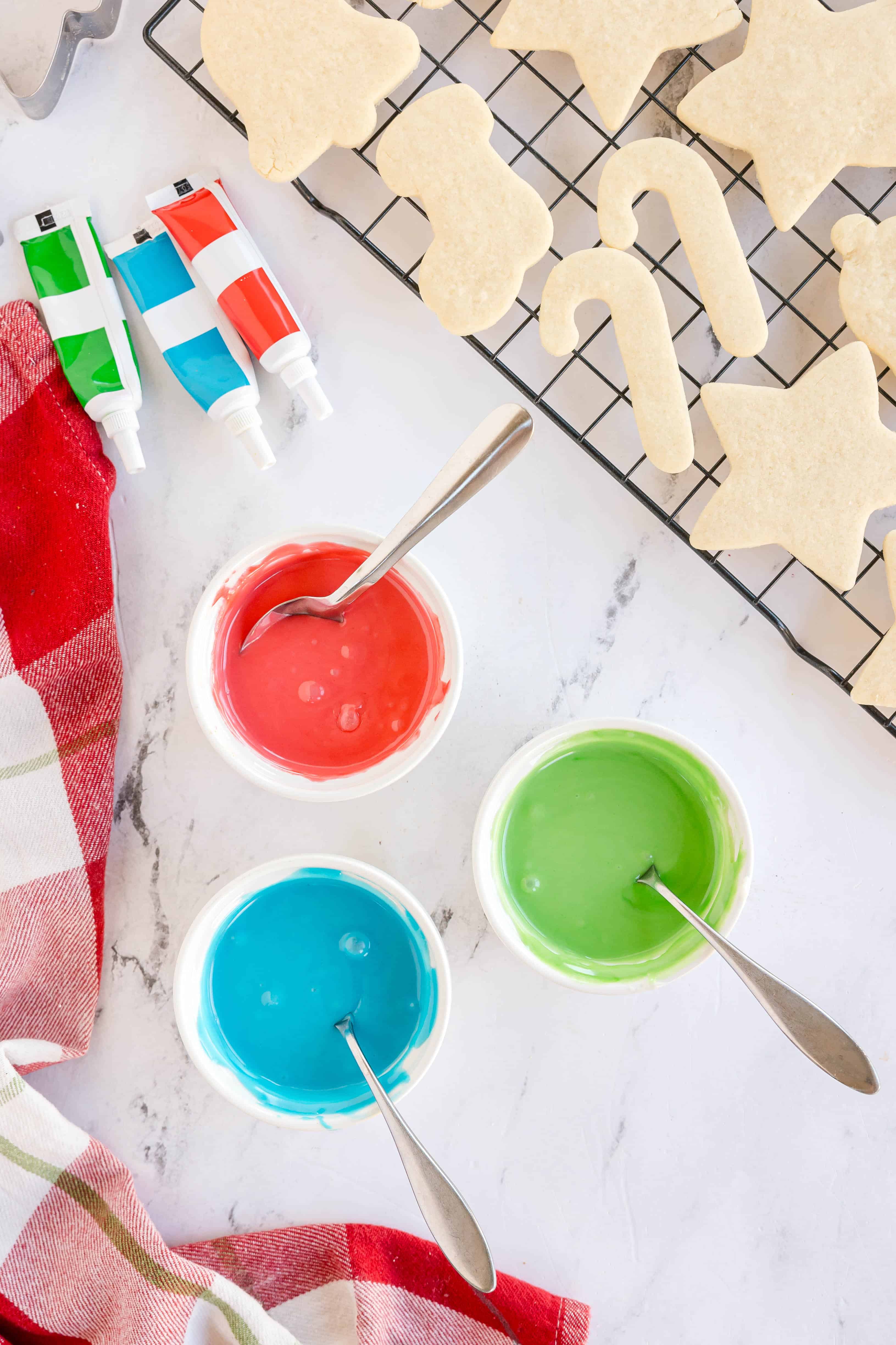 Three bowls of red, green, and blue icing with spoons, cookies on a cooling rack, and tubes of food coloring on a white surface.