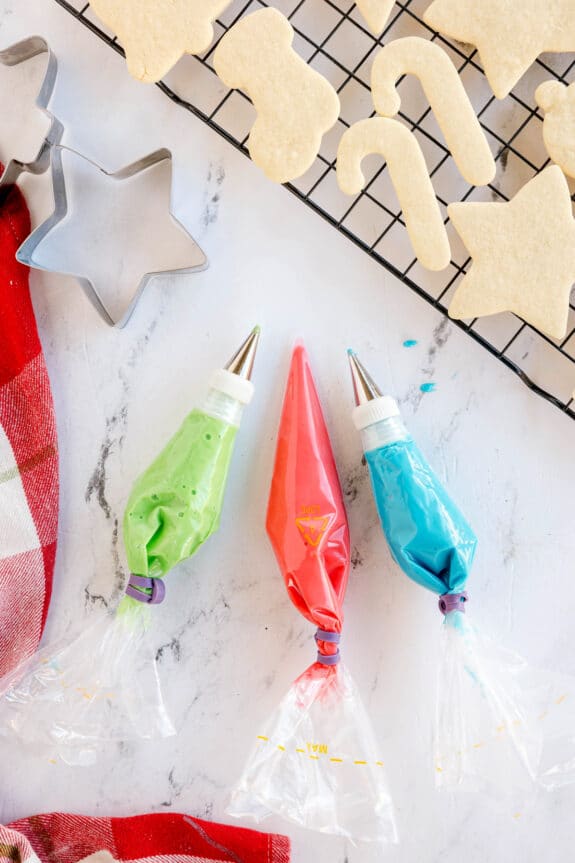 Next step in preparing Christmas Sugar Cookies is to make three piping bags with green, red, and blue icing. Each lie on a white surface next to a wire rack holding undecorated holiday cookies and metal cookie cutters.