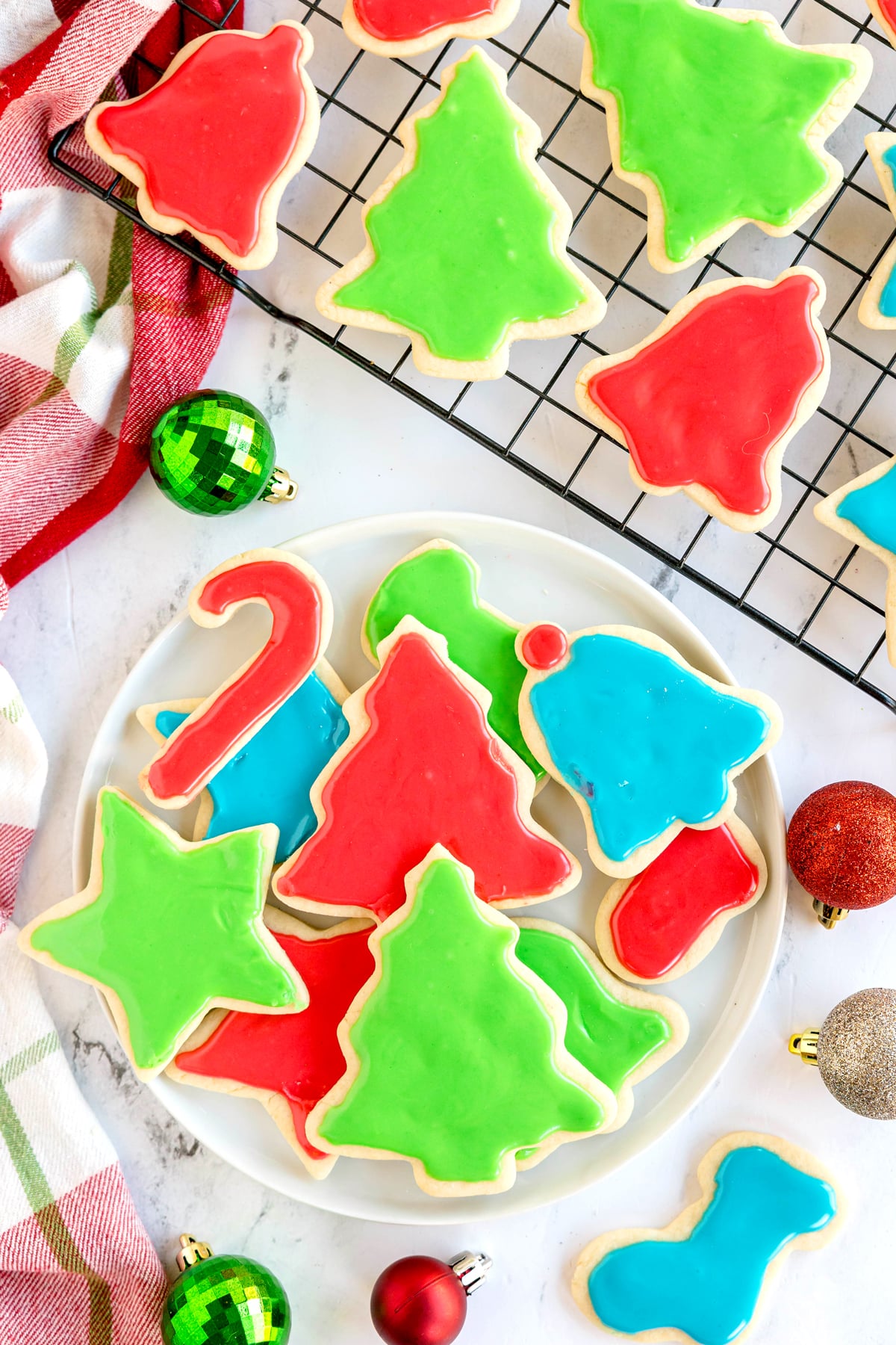 Christmas Sugar Cookies in holiday shapes and colors arranged on a plate and cooling rack, with ornaments and a striped towel nearby.