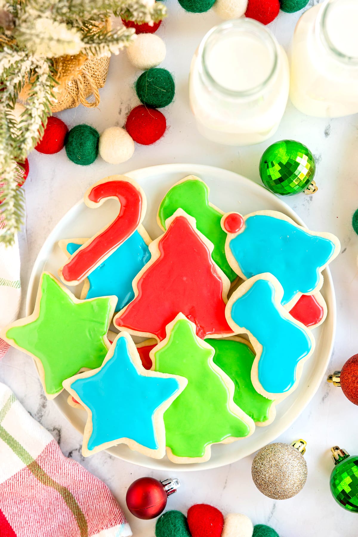 A plate of Christmas Sugar Cookies with colorful red, green, and blue icing, surrounded by ornaments and two bottles of milk.